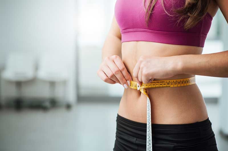 A fit woman in a pink sports bra and black workout leggings measuring her waist with a yellow tape measure, symbolizing weight loss progress. The image has a blue border with a checkmark in the top right corner.