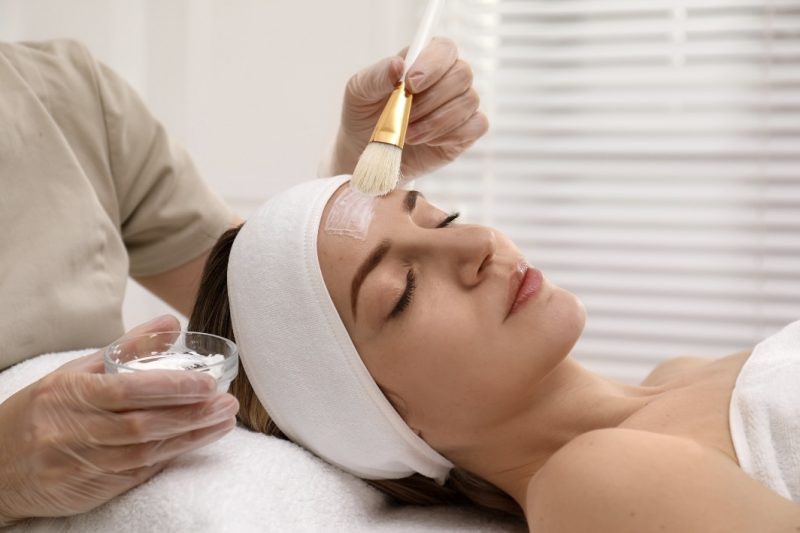 A woman reclining comfortably in a professional spa setting, receiving a chemical peel treatment by a skilled aesthetician. The aesthetician is carefully applying a peel solution to the woman’s face, focusing on rejuvenation and skin health.